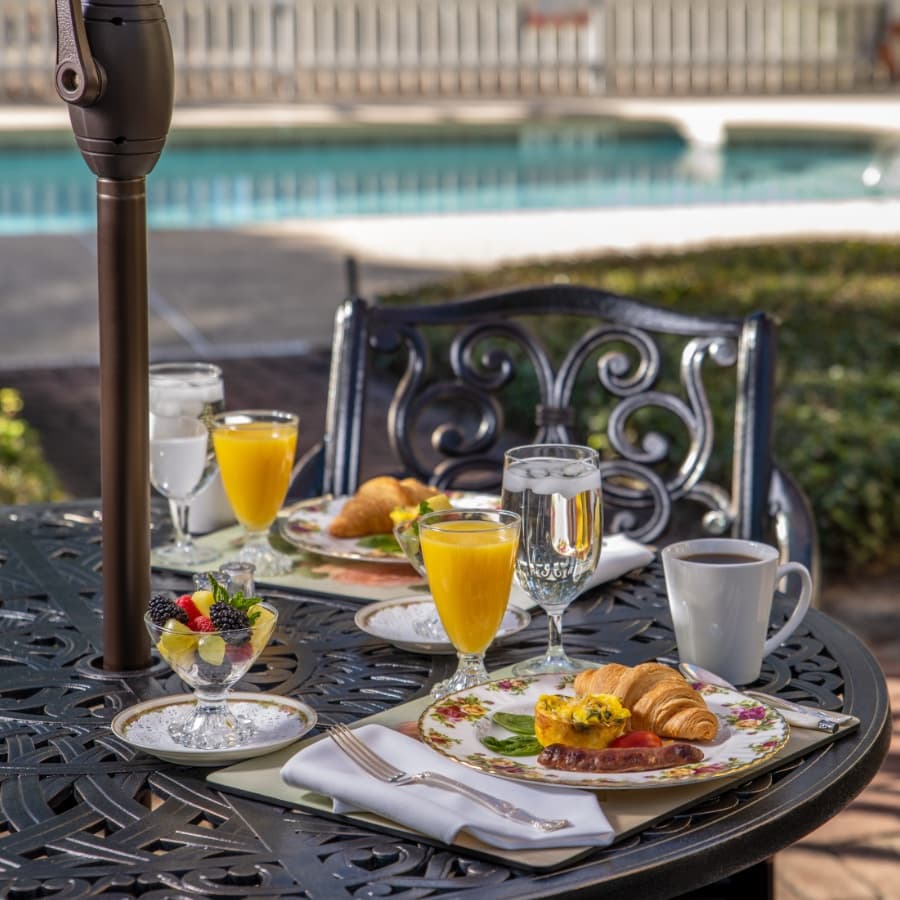A beautifully arranged outdoor breakfast table featuring plates of croissants, eggs, sausage, fruit, and drinks by a pool.
