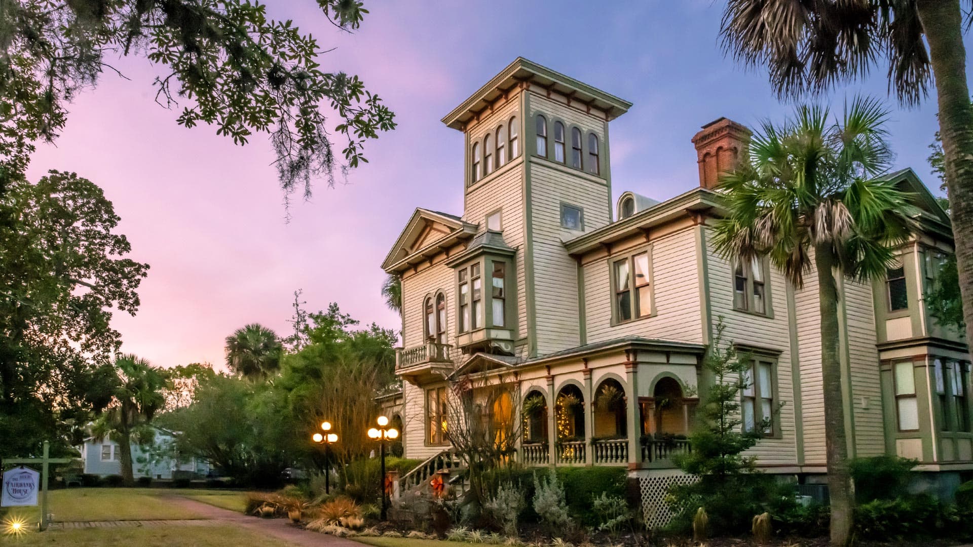 A historic Victorian-style house surrounded by palm trees at sunset.
