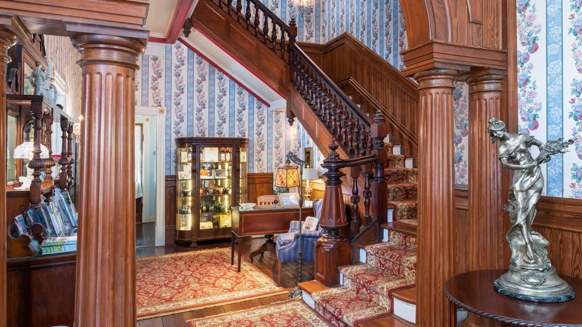 A grand staircase flanked by wooden columns, ornate wallpaper, and antique furnishings in a vintage foyer.