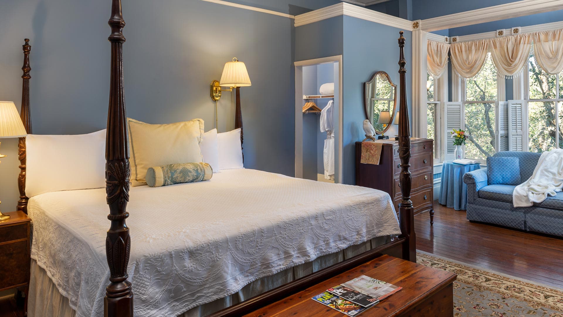 A cozy bedroom featuring a four-poster bed, a sofa, and large windows with natural light.