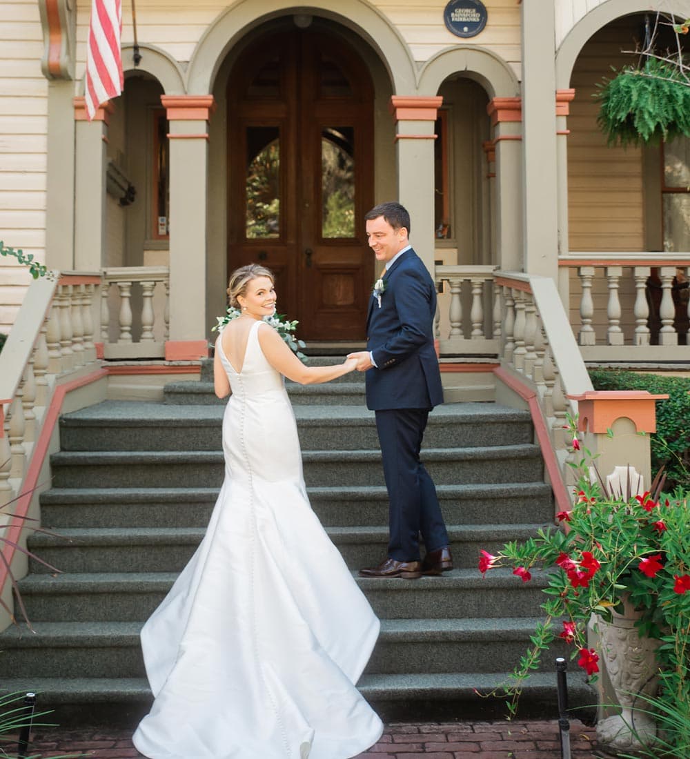 A bride and groom hold hands and smile at each other on the steps of a vintage house.