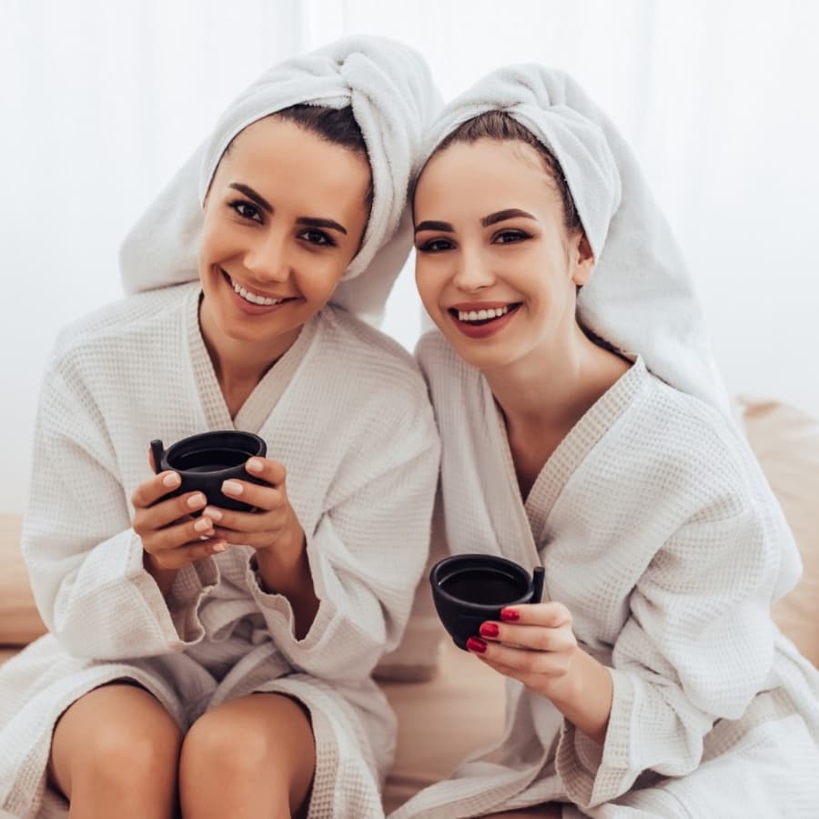 Two women in white bathrobes and towels smile while holding black cups.